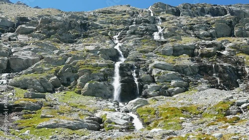 Waterfalls flowing in idyllic uncontaminated environment crossing green meadows and boulders on the Alps in summer. Slow motion