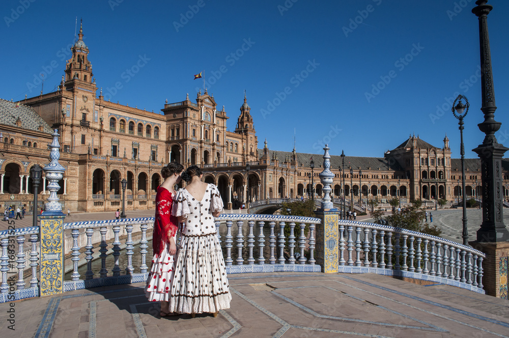 Fototapeta premium Spagna: ragazze sivigliane di spalle in abiti tipici in Plaza de Espana pronte per la Fiera di Siviglia (Feria de abril) che inizia due settimane dopo la Settimana Santa di Pasqua