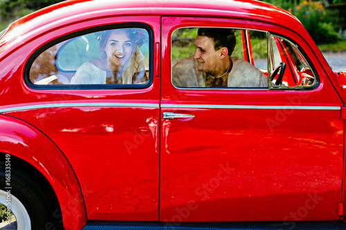 Beautiful happy young bride and groom in traditional clothes looking from red retro auto. Ukrainian wedding, culture, ethical traditions, concept.