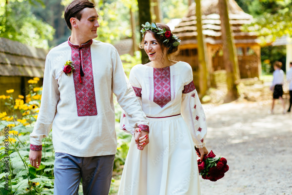 Beauty wedding couple in traditional clothes with red bouquet walking in old historical village. Ukrainian folklor wedding, culture, ethical traditions, concept.