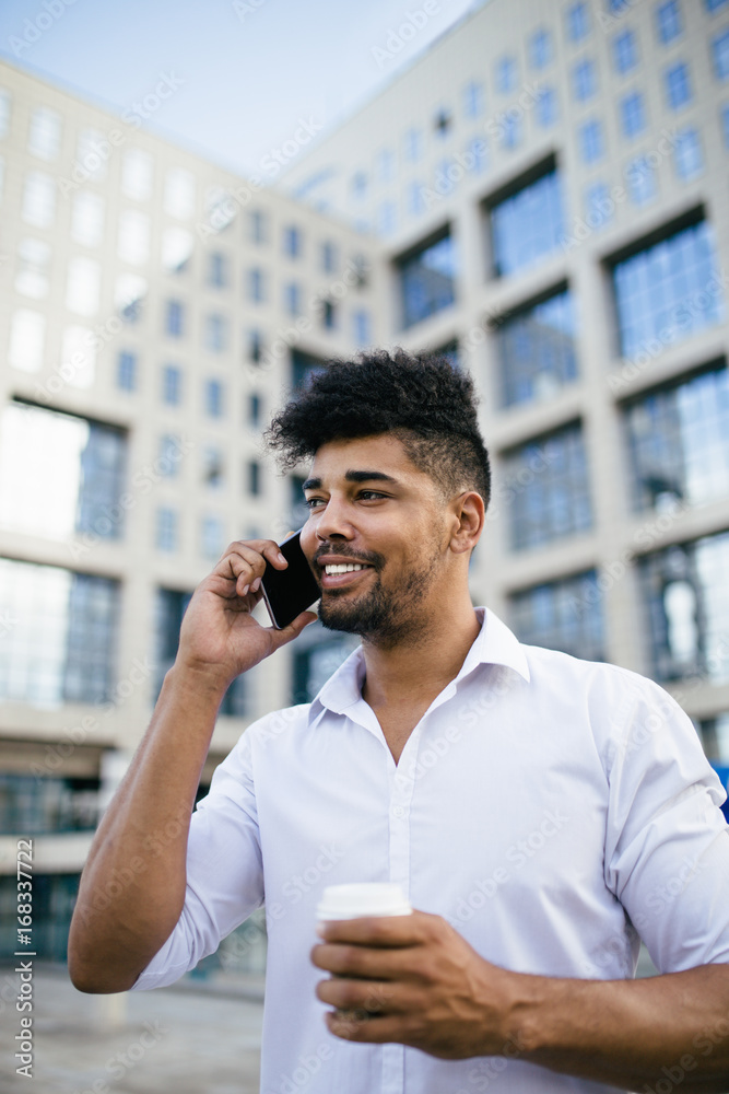 Fototapeta premium Young handsome Afro American man standing in front of huge modern business building smiling and talking on cell phone