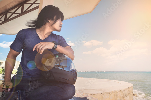 Foto An Asian man sitting under roof next to the sea with his helmet