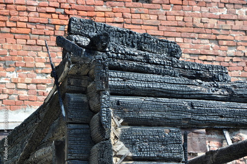 Close-up view of a charry corner of a burnt wooden house against a red brick wall.
 photo