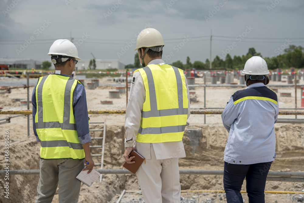 Group of workmen wearing protective helmets and vests walking among concrete walls of unfinished building showing development progress to foreman inspector on construction site Group of workmen wearing protective helmets and vests walking among concrete walls of unfinished building showing development progress to foreman inspector on construction site