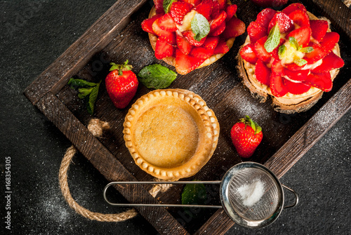 Summer and spring dessert. Home pies tartlets with custard and strawberries, decorated with mint and powdered sugar. On black stone table, rustic, with wooden board, tray. Copy space top view