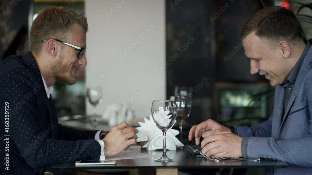 Two businessmen meets for discussion questions in their lunch in the cafe . Handsome young people sit at the table and talks about job. One of man holds pen in hands and then writes document on the Two businessmen meets for discussion questions in their lunch in the cafe . Handsome young people sit at the table and talks about job. One of man holds pen in hands and then writes document on the