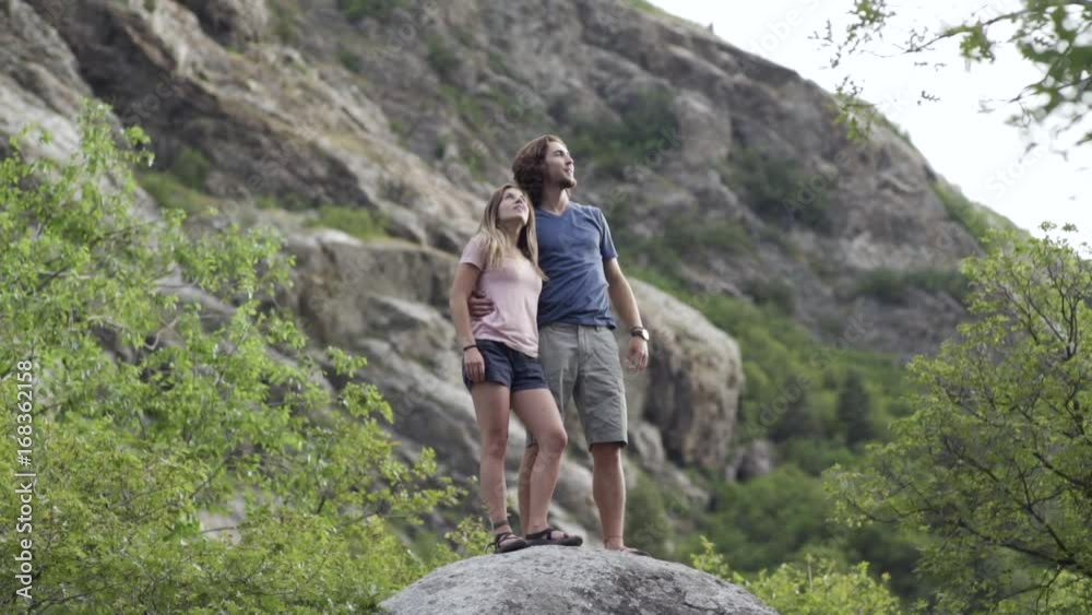 Couple Stand On Top Of Huge Boulder And Enjoy Mountain Views, They Turn, Point, Look, And Kiss Couple Stand On Top Of Huge Boulder And Enjoy Mountain Views, They Turn, Point, Look, And Kiss