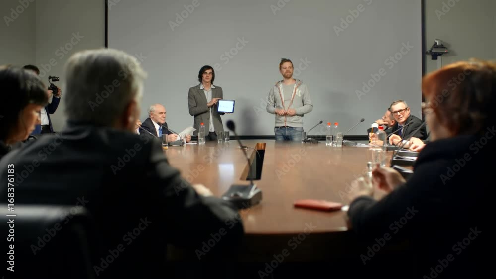 Large diverse group of businessmen appreciate the achievements of two young male partners from new start-up at the round table in modern conference hall of big company