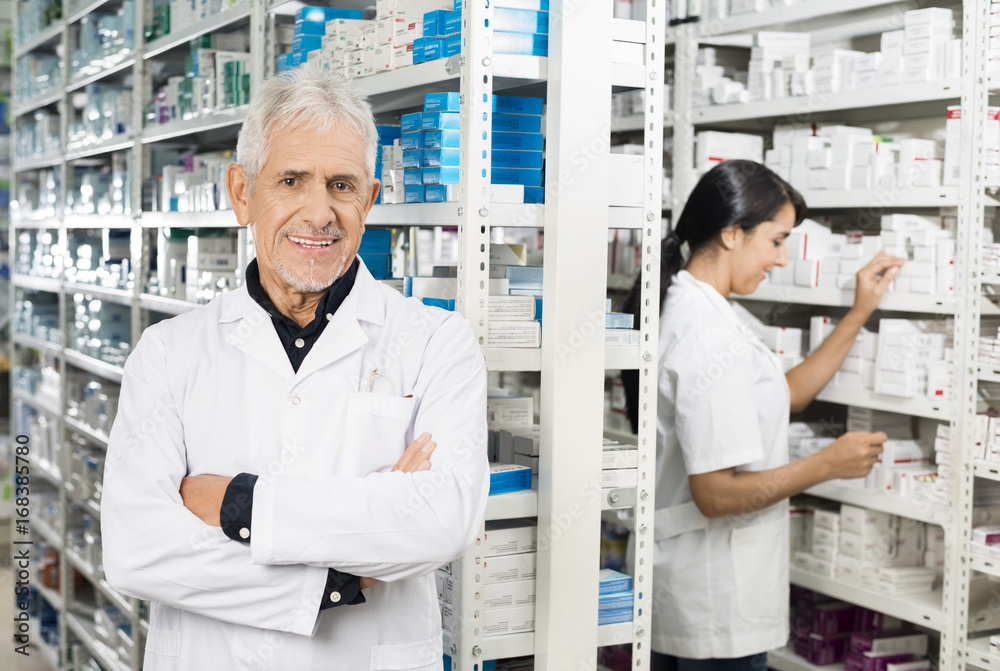 Senior Pharmacist Standing Arms Crossed While Colleague Checking