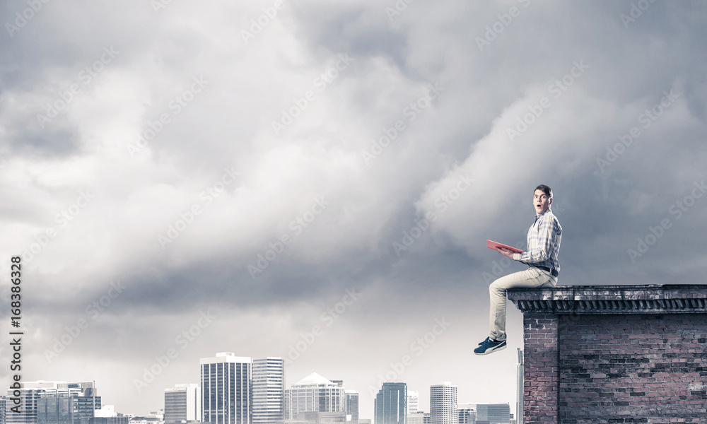 Man on roof edge reading book and cityscape at background