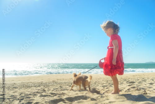 Little blond girl in red dress with dog on the beach