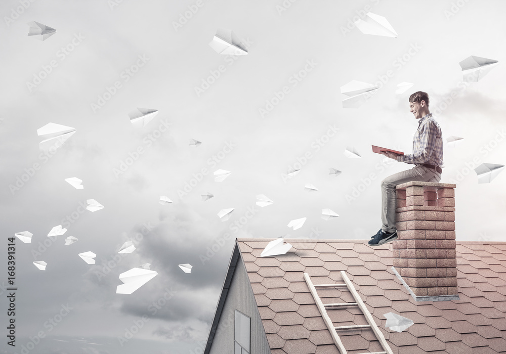 Man on brick roof reading book and paper planes flying in air Man on brick roof reading book and paper planes flying in air