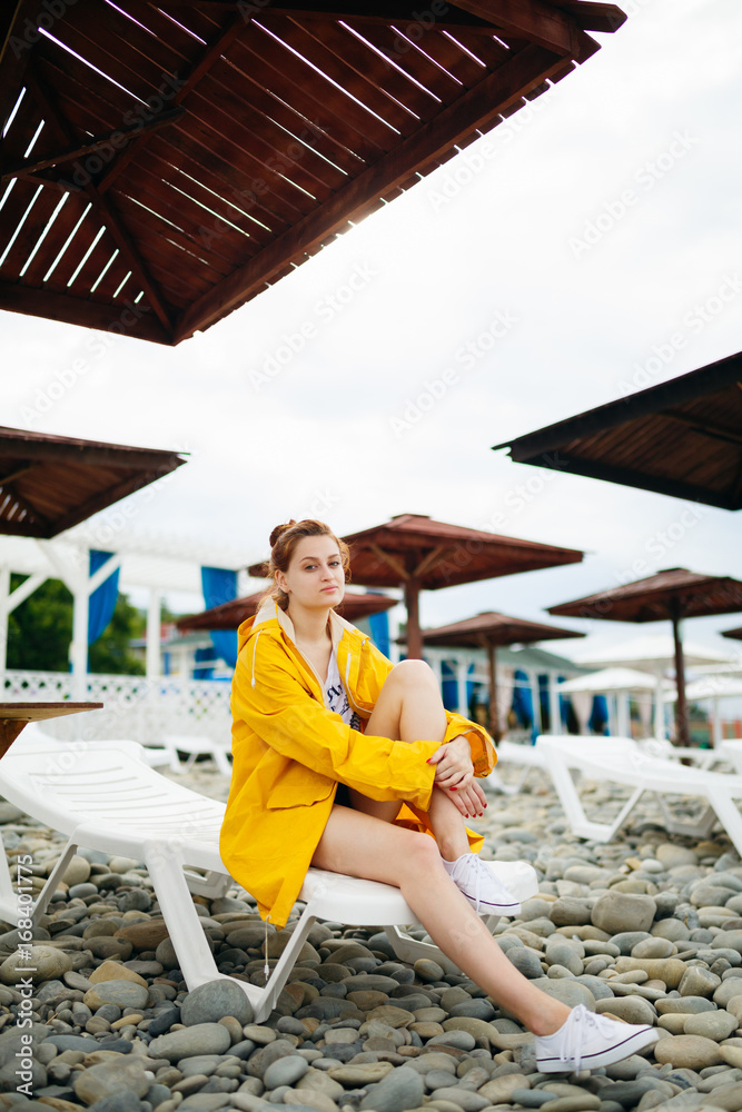 Young woman in bright raincoat sitting on sunbed on pebble beach looking at camera.