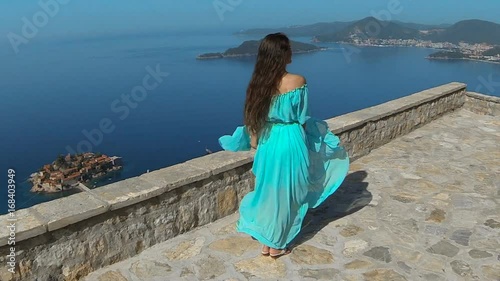 Beautiful brunette smiling Girl with Healthy Long Hair in blowing dress. Happy Young Woman Enjoying Nature and having Fun over sea and blue sky. Sveti Stefan, Montenegro.