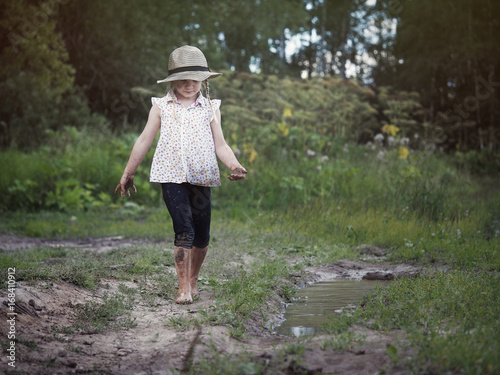 A child playing in a muddy puddle. Dirty girl in a hat and barefoot. Rural road © kozorog