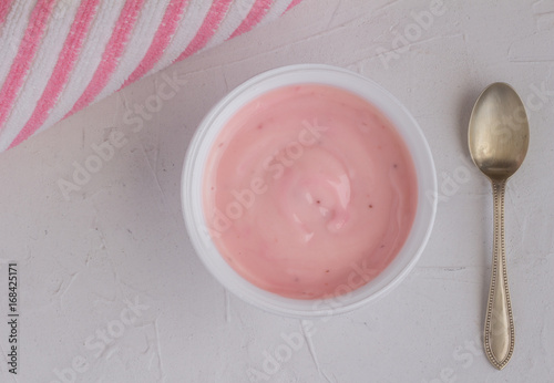 Healthy strawberry fruit flavored yogurt with natural coloring in plastic cup isolated on white background with little silver spoon and pink and white cloth - top view shot in studio