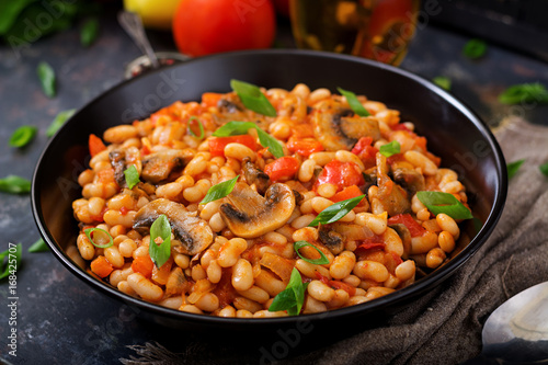 Stewed white beans with mushrooms and tomatoes with spicy sauce in a black bowl.
