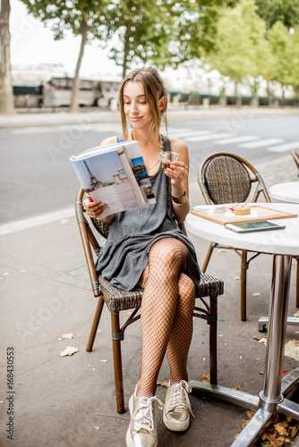 Young stylish woman reading journal sitting outdoors at the cafe in Paris