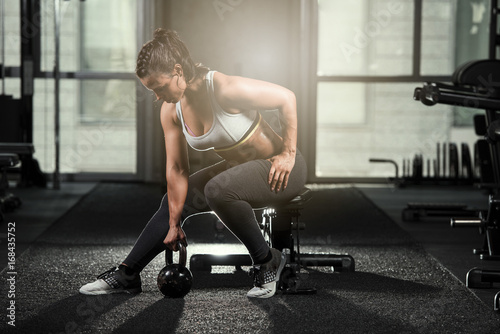 Strong brunette athletic woman sitting on a bench wearing a sports bra and black tights about to work out this a kettlebell in a dark gym with a light flair behind