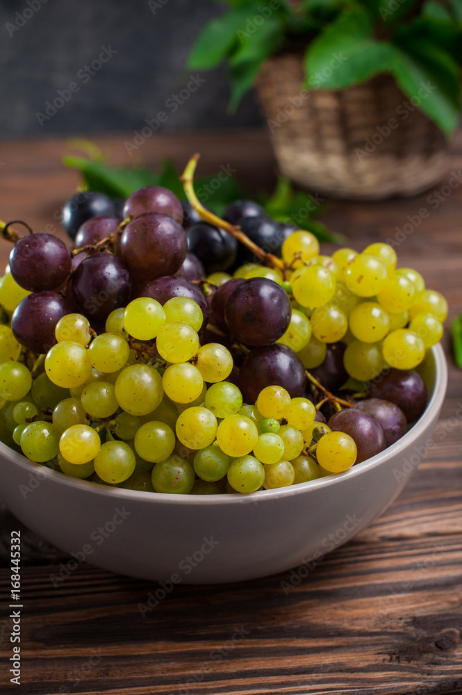 Close up bowl of various grapes: red, white and black berries on the dark wooden table with wicker basket with leaves in the background . Selective focus.