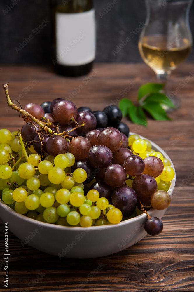 Close up bowl of various grapes: red, white and black berries on the dark wooden table with bottle and glass of white wine in the background . Selective focus. Close up bowl of various grapes: red, white and black berries on the dark wooden table with bottle and glass of white wine in the background . Selective focus.