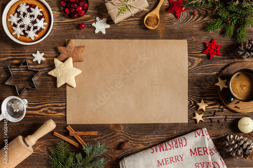 Christmass symbols sweets, candle, cinnamon, snowflakes and empty piece of paper on center. Christmass background with branches of furry spruce, cup of coffee on a wooden background.
