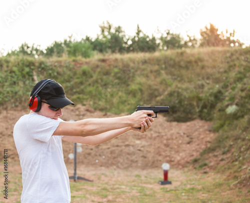 Handsome young policeman agent aiming and shooting at a target with a pistol desert eagle gun wearing black hat and white shirt in nature ambient firing firearms bullets in the air training