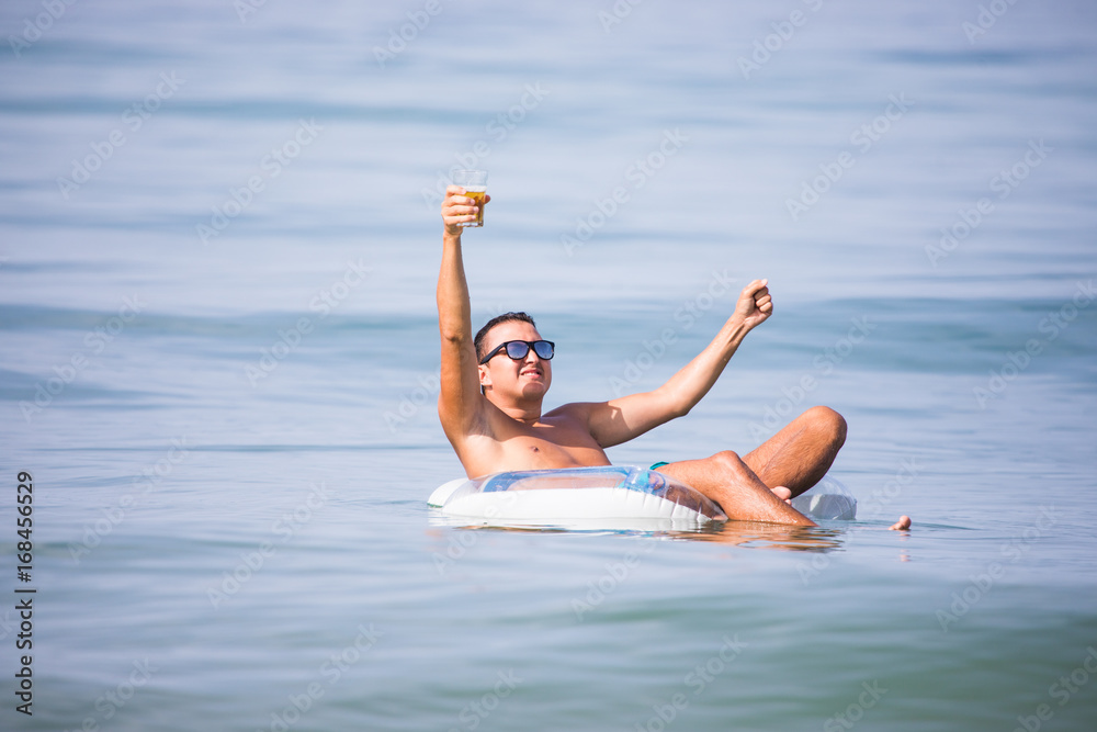 Young man with hands up in sunglasses with a glass of beer floating on rubber ring in the ocean water at sunset
