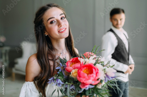 Close-up portrait of cheerful, young bride with a wedding bouquet on blurred groom background. A happy girl posing with a bunch of flowers
