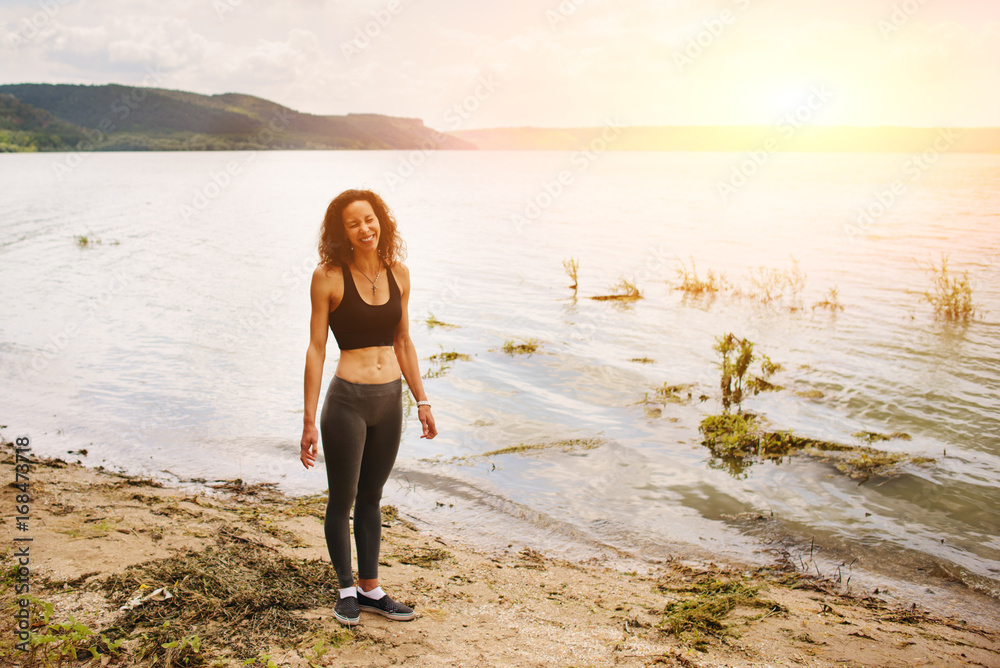 A beautiful sporty woman standing on the shore of a lake in sportswear and preparing for an exercising