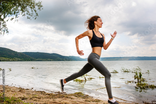 A beautiful sporty woman runing on the shore of a lake in sportswear. Girl is exercising