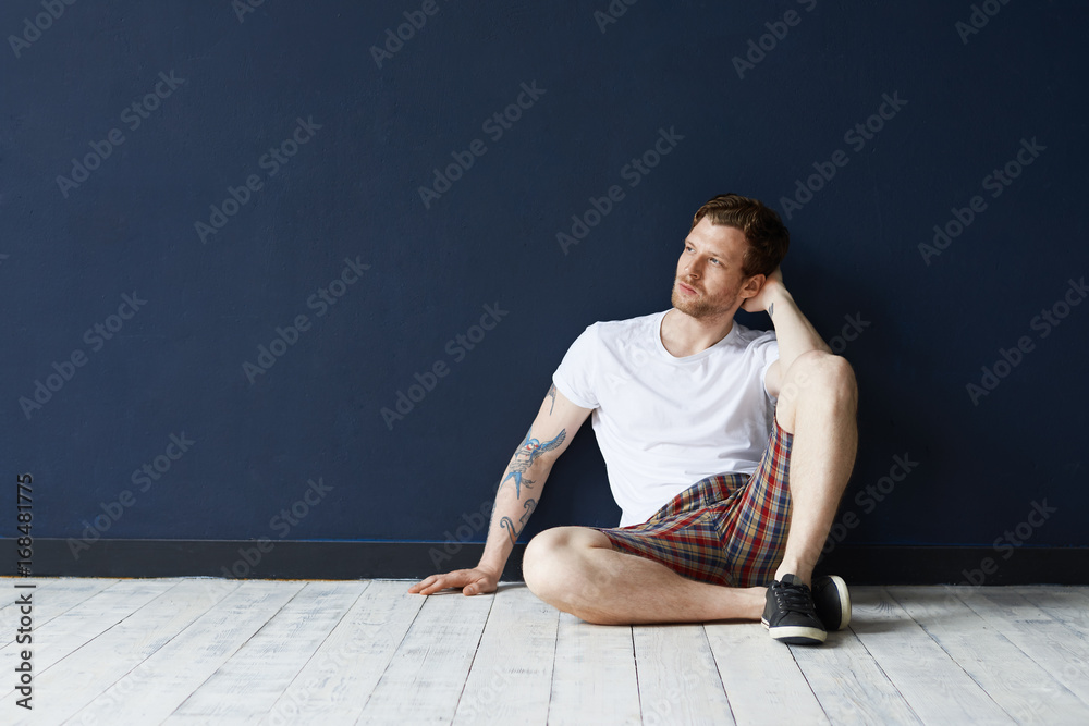 Fototapeta premium Picture of attractive young unshaven Caucasian man with tattoos wearing black sneakers, t-shirt and shorts having rest indoors, thinking about his life, future plans, looking thoughtful or dreamy
