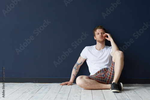 Indoor shot of handsome bearded young tattooed male of European appearance relaxing at home, sitting on floor wearing stylish clothes. People, lifestyle, rest, leisure and relaxation concept