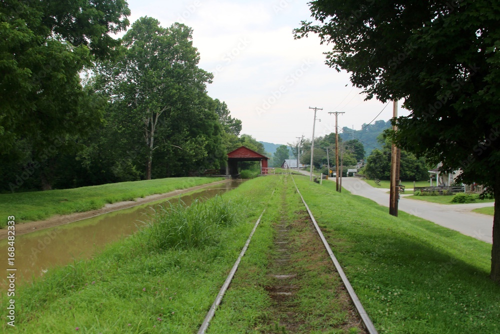 The railroad tracks in the tall grass by the canal and past the covered bridge.