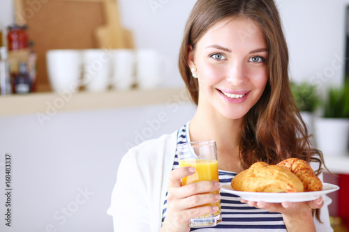 Young woman with glass of juice and cakes standing in kitchen