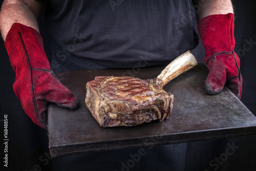 Man is holding barbecue wagyu Tomahawk steak on old board in his hands with gloves