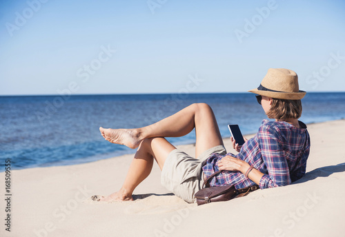 Young woman lying on a beach with smart phone. Technology and travel concept. People using mobile devices to stay connected from remote parts of the world