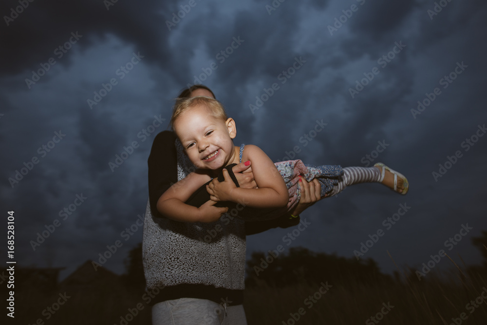 Cute baby girl playing with her mother outdoor. Flying against dark sky. Cute baby girl playing with her mother outdoor. Flying against dark sky.