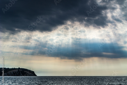 Sun rays on dark cloudy sky with coast in the background. © jakazvan