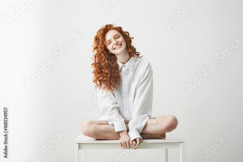 Tender young redhead girl in shirt smiling looking at camera sitting on table posing over white background. photo