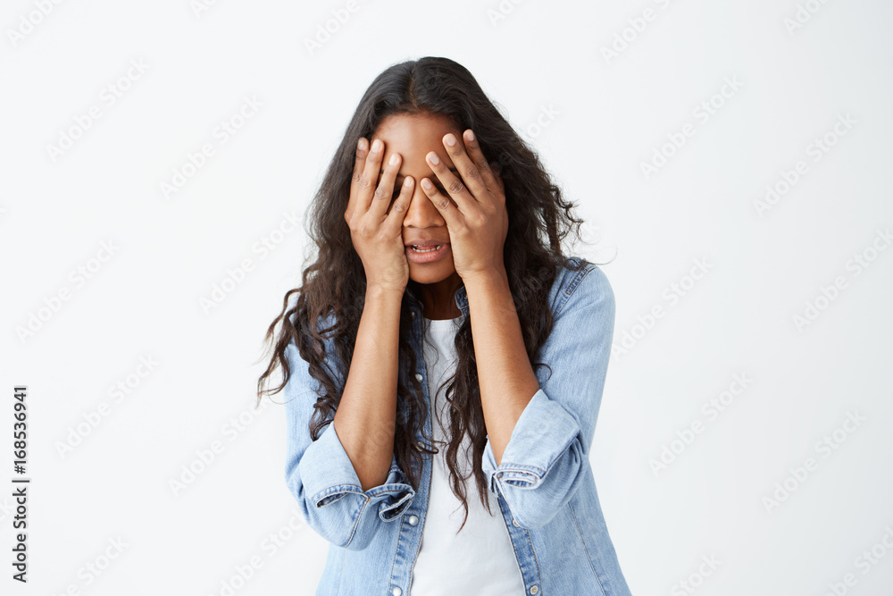 Stressed Afro-American female with long wavy hair casually dressed feeling upset, keeping hands on her head, hiding her face in despair after she heard bad news. Stressed Afro-American female with long wavy hair casually dressed feeling upset, keeping hands on her head, hiding her face in despair after she heard bad news.