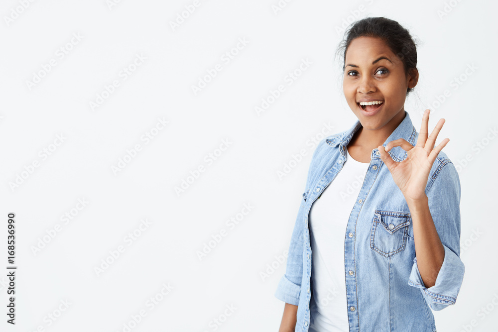 Happy satisfied Afro-american female wearing denim shirt making okay sign and smiling cheerfully, showing her support and respect to someone. Positive emotions concept. Happy satisfied Afro-american female wearing denim shirt making okay sign and smiling cheerfully, showing her support and respect to someone. Positive emotions concept.