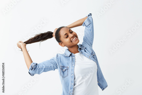 Indoor shot of Afro-American girl with long wavy hair gathered in ponytail and dark skin dressed in denim shirt posing indoors, playing with her hair. photo