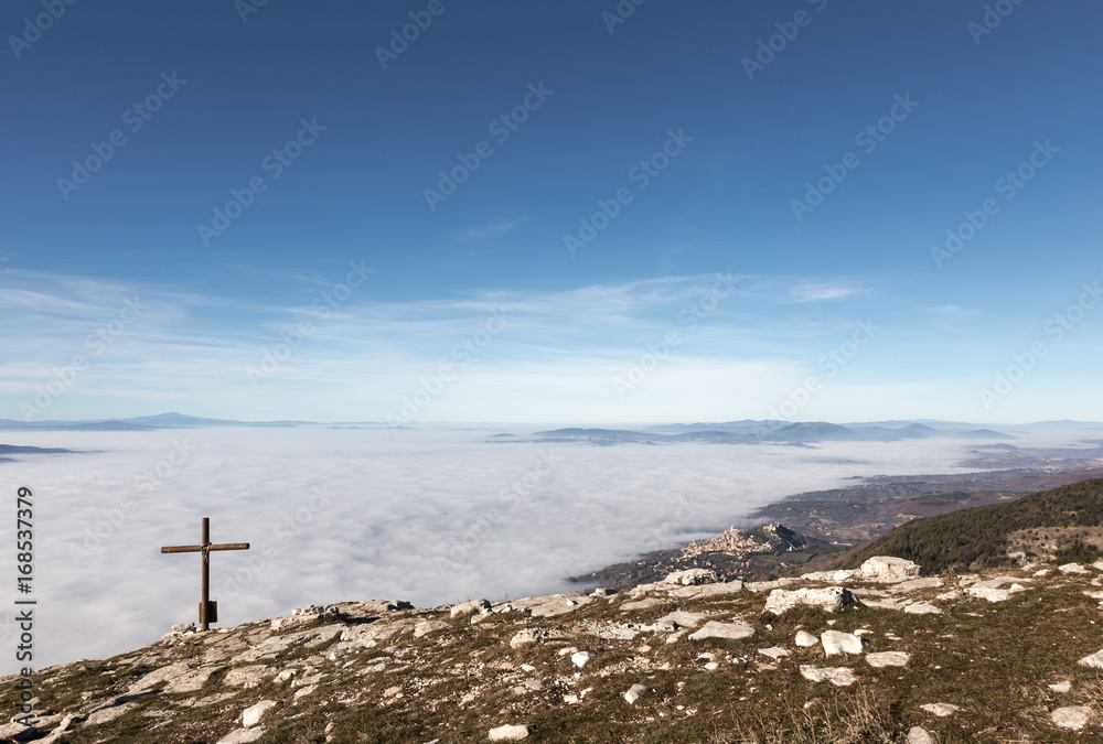 A wooden cross on top of Subasio mountain, with a sea of fog below and Assisi town (Umbria) in the background