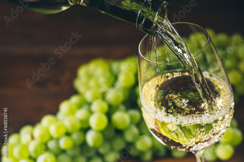 Pouring white wine into a glass with a bunch of green grapes against wooden background photo