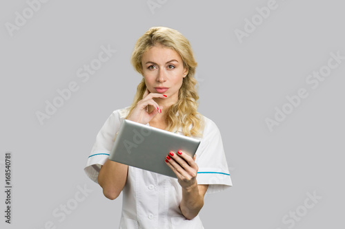 Portrait of young nurse with digital tablet. Thoughtful young female doctor or nurse holding computer tablet and having an idea, grey background. © DenisProduction.com