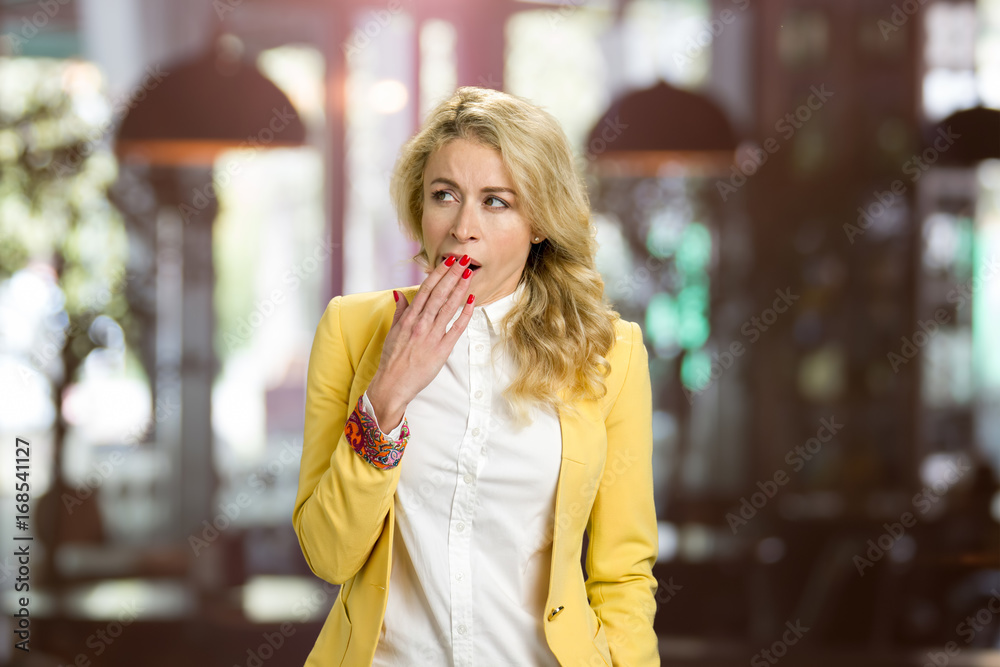 Fototapeta premium Young yawning woman looking away. Closeup portrait young blond woman placing hand covering mouth, yawning, looking away bored on blurred background.