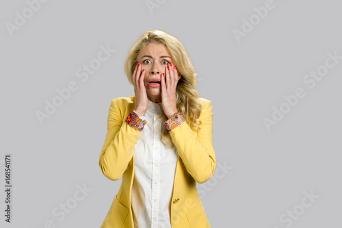 Portrait of horrified young woman. Desperate young blond woman touching her face looking confused and terrified, grey background.