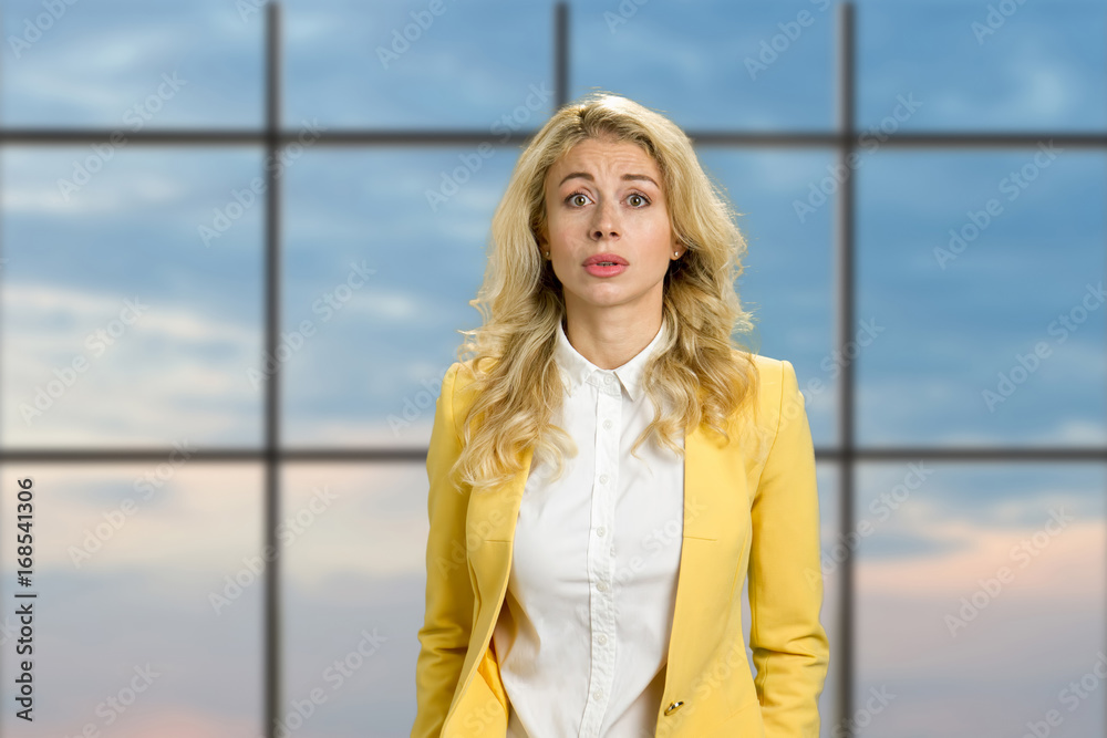 Fototapeta premium Portrait of disappointed young woman. Beautiful upset blonde looking confused on blue sky office window background. Human facial expressions.
