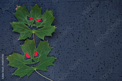 Dark halloween background - a faces on an green maple leaves with red eyes. Copy space